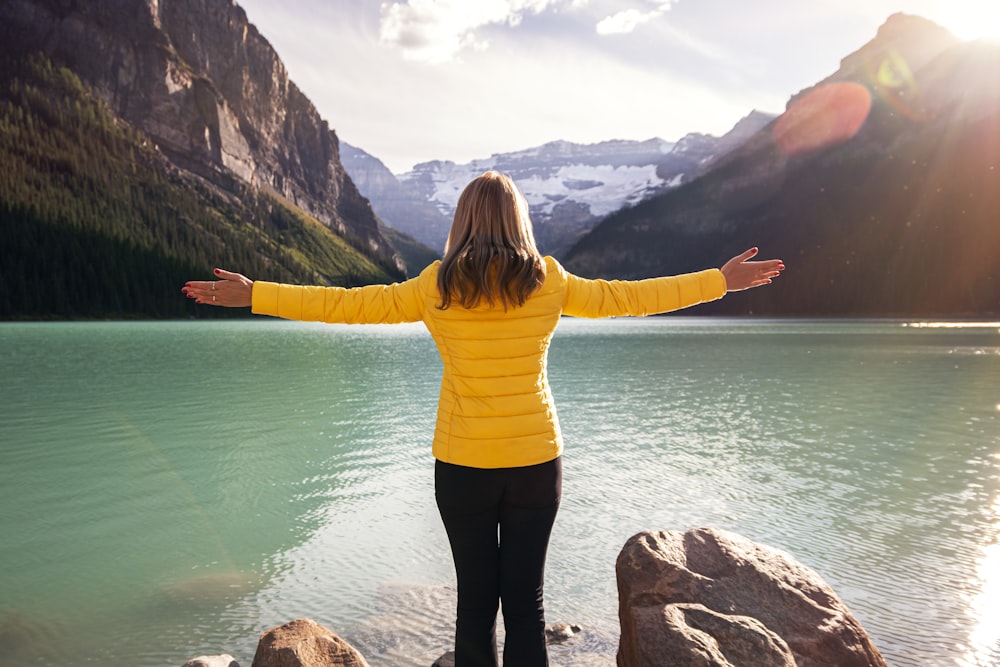 woman in yellow long sleeve shirt and black pants standing on rock near body of water