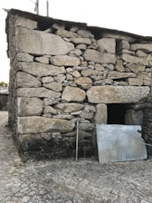 A sturdy structure made of large, irregular stones stacked to form a wall. A metal panel leans against the building next to a steel pipe. The ground is covered with cobblestone pavement, and the building appears weathered, suggesting age.