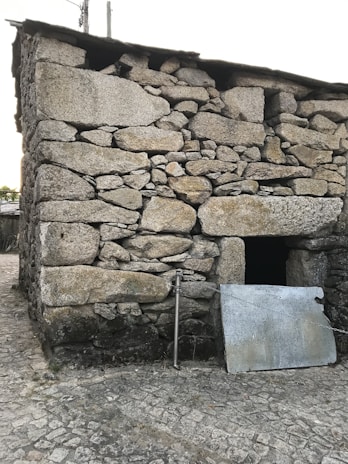 A sturdy structure made of large, irregular stones stacked to form a wall. A metal panel leans against the building next to a steel pipe. The ground is covered with cobblestone pavement, and the building appears weathered, suggesting age.