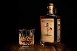 A bottle of Barterhouse Kentucky Bourbon along with a crystal glass containing a small amount of the whiskey is placed on a wooden surface. The bottle's label is clearly visible, featuring a vintage-style design. The background is dark, enhancing the focus on the bottle and glass.