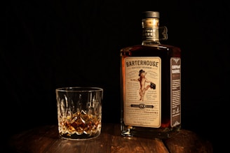 A bottle of Barterhouse Kentucky Bourbon along with a crystal glass containing a small amount of the whiskey is placed on a wooden surface. The bottle's label is clearly visible, featuring a vintage-style design. The background is dark, enhancing the focus on the bottle and glass.