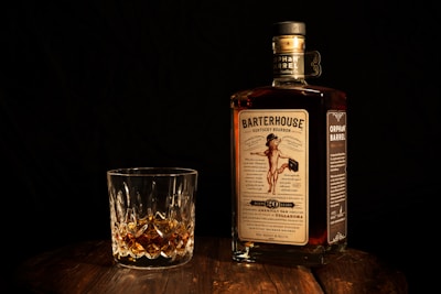 A bottle of Barterhouse Kentucky Bourbon along with a crystal glass containing a small amount of the whiskey is placed on a wooden surface. The bottle's label is clearly visible, featuring a vintage-style design. The background is dark, enhancing the focus on the bottle and glass.