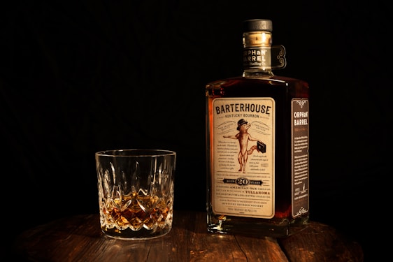 A bottle of Barterhouse Kentucky Bourbon along with a crystal glass containing a small amount of the whiskey is placed on a wooden surface. The bottle's label is clearly visible, featuring a vintage-style design. The background is dark, enhancing the focus on the bottle and glass.