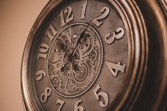 An ornate clock with exposed mechanical gears, surrounded by large, bold numbers. The clock has a vintage aesthetic, with a metallic finish that resembles bronze or brass. The gears are intricately designed and visible through the clock face, adding a steampunk element.
