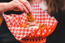 A hand holds a piece of crispy fried chicken being dipped into a small cup of white sauce. The chicken is placed in a brightly colored red basket lined with red and white checkered paper. The setting suggests a casual dining environment.