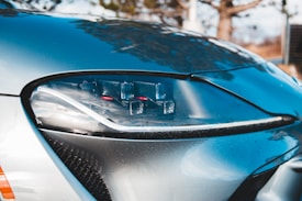 A close-up view of a modern car's headlight with a sleek design, featuring LED elements and a glossy finish. The car's body reflects the surrounding environment, adding a dynamic and polished appearance.