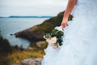 A bride holding a bouquet of pale pink and cream roses, standing on a grassy cliffside overlooking the ocean. The wedding dress is adorned with delicate embellishments and ruffles, creating a flowing and elegant appearance.