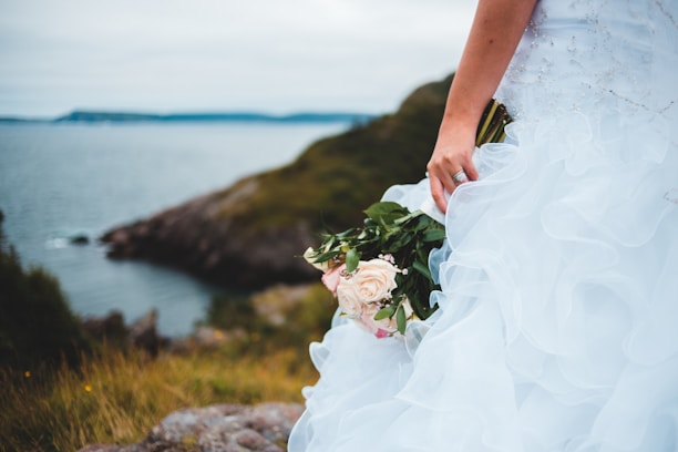 A bride holding a bouquet of pale pink and cream roses, standing on a grassy cliffside overlooking the ocean. The wedding dress is adorned with delicate embellishments and ruffles, creating a flowing and elegant appearance.