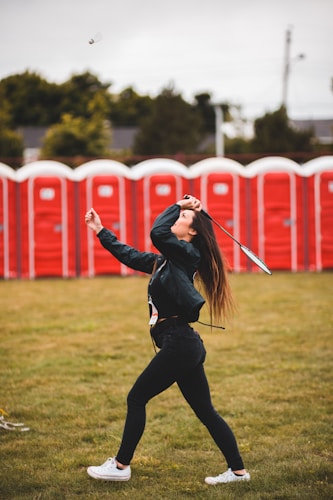 A person is playing badminton in a grassy area with a series of red portable toilets visible in the background. The individual is mid-swing, focusing on hitting a shuttlecock, wearing casual clothing including jeans and a jacket.
