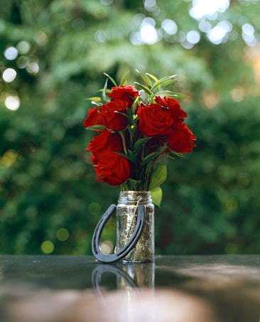 A bouquet of vibrant red roses with lush green leaves is arranged in a rustic, reflective jar. A horseshoe is attached to the front of the jar, adding a rustic charm. The background features a soft-focus, natural green setting.