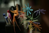 A close-up of colorful origami flowers crafted from paper straws, with a blurred background of pens, markers, and scissors in a holder.