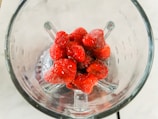 A group of frozen strawberries is placed inside a glass blender jar, viewed from above. The berries have a frosty appearance, indicating they are frozen. The clear glass of the blender allows a full view of the strawberries sitting at the bottom.