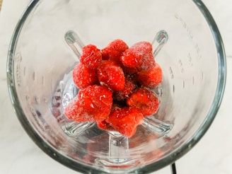 A group of frozen strawberries is placed inside a glass blender jar, viewed from above. The berries have a frosty appearance, indicating they are frozen. The clear glass of the blender allows a full view of the strawberries sitting at the bottom.