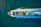 Aerial view of a large cargo ship traversing through the ocean, carrying colorful shipping containers on its deck. The vessel creates noticeable waves in the deep blue water, leaving a wake behind.