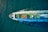 Aerial view of a large cargo ship traversing through the ocean, carrying colorful shipping containers on its deck. The vessel creates noticeable waves in the deep blue water, leaving a wake behind.