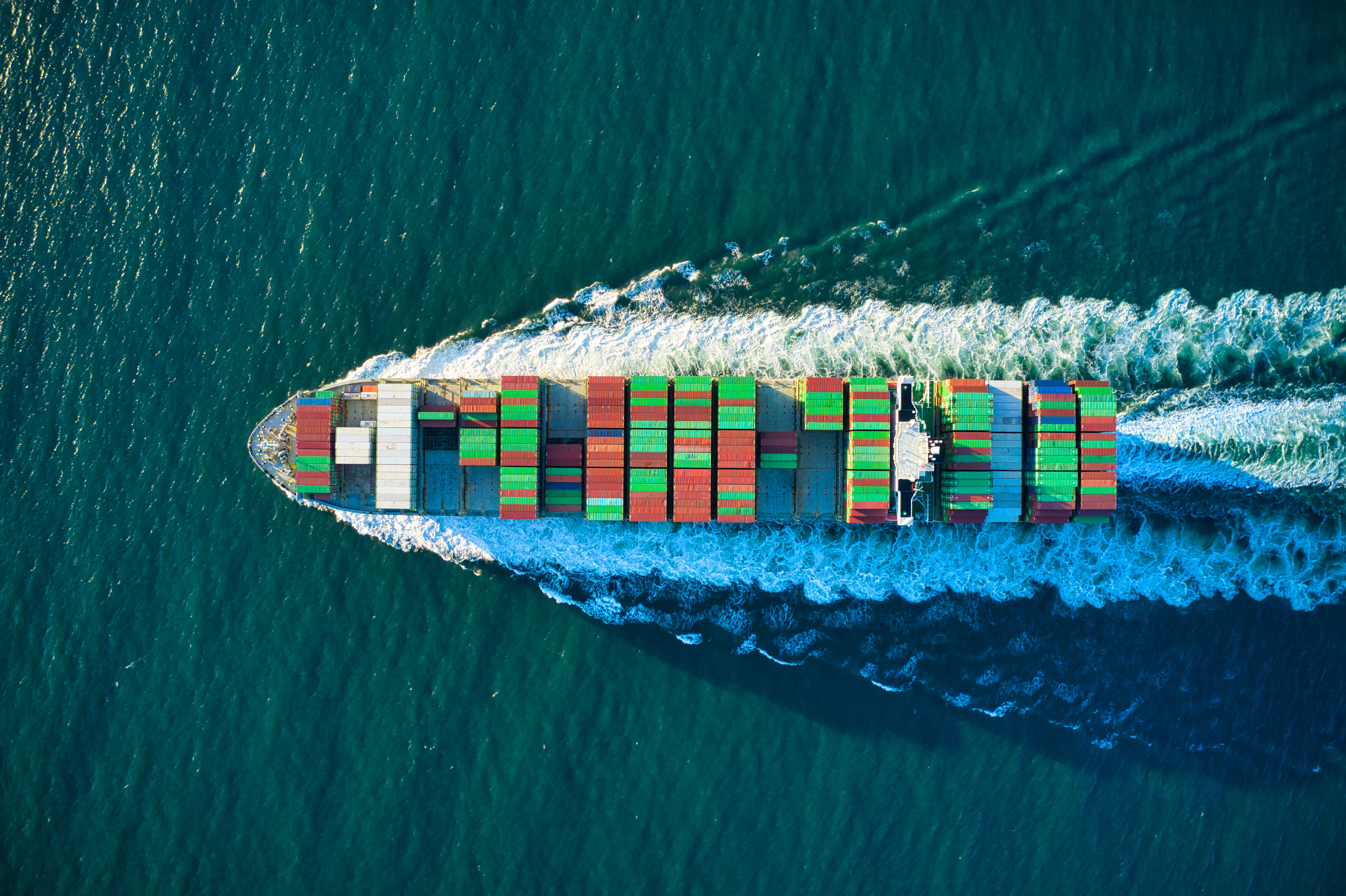A fully-laden container ship photographed from directly above, cutting a white wake through deep green water.