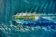 Aerial view of a large cargo or tanker ship moving through the ocean, creating waves and ripples in the surrounding water. The ship is green with yellow markings and appears to be cutting through deep blue waters. White foamy trails are visible behind the ship due to its movement.