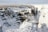 A snow-covered canyon with rugged rock formations and a lightly snow-dusted landscape in the background. The rock faces are steep, showcasing a mix of gray and white tones, with patches of exposed rock contrasting against the snow.