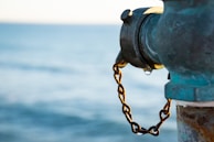 A close-up of a weathered fire hydrant with a rusty chain attached. The background features a serene, blurred body of water, suggesting a location near a waterfront.