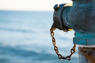 A close-up of a weathered fire hydrant with a rusty chain attached. The background features a serene, blurred body of water, suggesting a location near a waterfront.