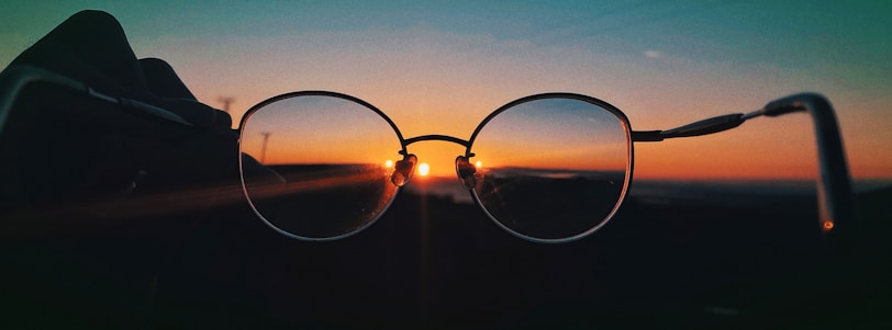 A pair of round glasses is held up against a backdrop of a sunset. The sun is positioned almost centrally within the lenses, creating a striking silhouette and casting warm hues across the frame.