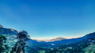 Panoramic view from a hilltop overlooking lush green valleys under a bright blue sky.