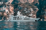 A luxurious yacht is floating on calm blue waters with a stunning backdrop of rugged cliffs and a hillside covered with quaint Mediterranean houses. The sky is clear, enhancing the serene and picturesque landscape.