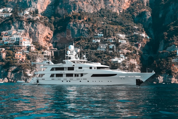 A luxurious yacht is floating on calm blue waters with a stunning backdrop of rugged cliffs and a hillside covered with quaint Mediterranean houses. The sky is clear, enhancing the serene and picturesque landscape.