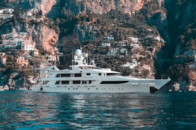 A luxurious yacht is floating on calm blue waters with a stunning backdrop of rugged cliffs and a hillside covered with quaint Mediterranean houses. The sky is clear, enhancing the serene and picturesque landscape.