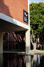 A modern building with a brick facade labeled 'McIntosh Humanities Building.' It has large concrete pillars and a reflective pool of water in the foreground. Tall green trees provide shade, and the scene is calm and serene.