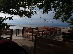 A serene beach setting with wooden tables and chairs on a concrete patio. Overhead, string lights are hung between lush green branches of trees framing the scene. The ocean is visible in the background with several boats anchored near the shoreline. The sky is partly cloudy with the sun casting soft shadows.
