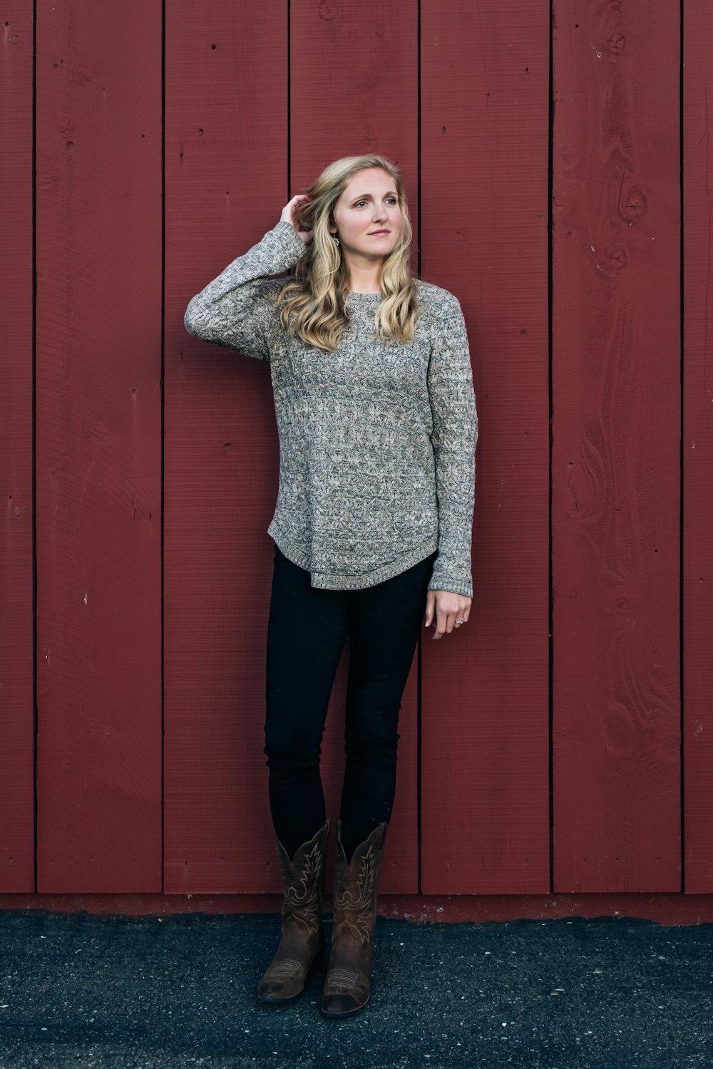 woman in gray sweater and black pants standing beside red wooden wall