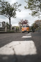 A vintage yellow van with a distinctive logo is parked on a quiet street lined with trees and stone fences. The perspective emphasizes the road leading towards the vehicle, with white dashed lines in the foreground and lush green foliage overhead. The sky is overcast, adding a sense of calm to the setting.