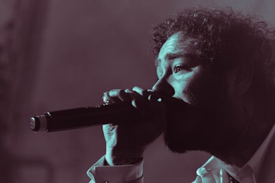 A person is singing passionately into a microphone. The image is in a low-light setting with a focus on the face and microphone. The person's curly hair, facial features, and expression convey intensity and emotion.