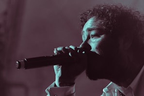 A person is singing passionately into a microphone. The image is in a low-light setting with a focus on the face and microphone. The person's curly hair, facial features, and expression convey intensity and emotion.