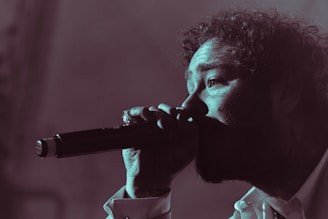 A person is singing passionately into a microphone. The image is in a low-light setting with a focus on the face and microphone. The person's curly hair, facial features, and expression convey intensity and emotion.