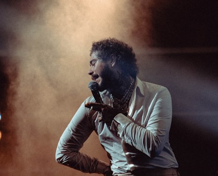A performer holds a microphone, dressed in a light-colored shirt under dramatic stage lighting with smoke effects. The atmosphere appears dynamic and engaging.