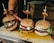 Three gourmet burgers are arranged on a wooden board. The buns are sprinkled with sesame seeds and have skewers topped with grilled meat and a pickled onion. The burgers are filled with beef patties, fresh arugula, tomato slices, and a dollop of creamy cheese.