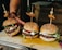 Three gourmet burgers are arranged on a wooden board. The buns are sprinkled with sesame seeds and have skewers topped with grilled meat and a pickled onion. The burgers are filled with beef patties, fresh arugula, tomato slices, and a dollop of creamy cheese.