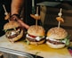 Three gourmet burgers are arranged on a wooden board. The buns are sprinkled with sesame seeds and have skewers topped with grilled meat and a pickled onion. The burgers are filled with beef patties, fresh arugula, tomato slices, and a dollop of creamy cheese.