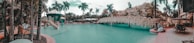 A tropical pool setting with palm trees surrounding a lagoon-style pool. There is a water slide leading into the pool, and a rock formation with sculptures resembling animals in the background. Sun loungers are placed around the pool area, indicating a leisure-focused environment.