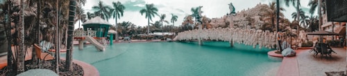 A tropical pool setting with palm trees surrounding a lagoon-style pool. There is a water slide leading into the pool, and a rock formation with sculptures resembling animals in the background. Sun loungers are placed around the pool area, indicating a leisure-focused environment.