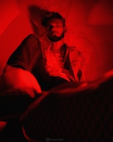 A person is seated in a dimly lit environment with a red hue. The person appears relaxed, wearing casual clothing, and is highlighted by dramatic lighting that emphasizes shadows and contours. The use of red lighting creates a surreal and artistic effect.