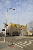 A street with a traffic light and traffic signs at an intersection. An off-white building with several windows stands in the background. Steam is rising from a manhole in the middle of the road. A tall communication tower is visible behind the building. Trees and street signs are present along the sidewalk.