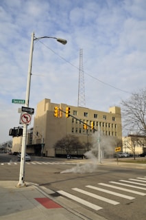 A street with a traffic light and traffic signs at an intersection. An off-white building with several windows stands in the background. Steam is rising from a manhole in the middle of the road. A tall communication tower is visible behind the building. Trees and street signs are present along the sidewalk.