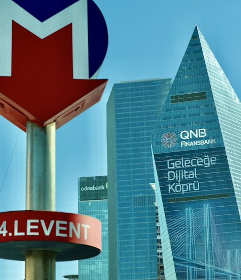 A modern urban landscape featuring tall skyscrapers with reflective glass facades. Prominent signage includes QNB Finansbank and Odeabank logos. A subway station sign labeled '4. Levent' is in the foreground, characterized by a red and blue arrow symbol.