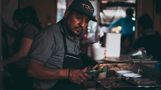 A man in a cap and striped shirt is working at a food stall, preparing a dish using tongs. He is surrounded by various cooking utensils and food containers. The setting appears to be indoors with a dimly lit ambiance, and there are other people in the background.