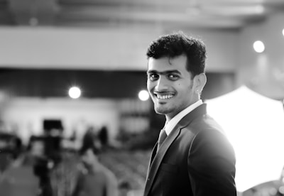 A person dressed in formal attire, wearing a suit and tie, smiles confidently while standing in a well-lit indoor setting. The background is softly focused, highlighting lights and blurred figures of other people.
