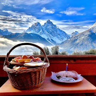 A picturesque landscape featuring snow-capped mountains under a vibrant blue sky with scattered clouds. The foreground shows a wooden table with a wicker basket filled with breakfast items such as cheese, eggs, and ham. Beside it, there is a small cake with a lit candle, placed on a doily.