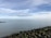 A calm seascape with an expansive body of water extending to the horizon under a partly cloudy sky. A line of distant wind turbines is visible on the horizon. In the foreground, a rocky shoreline is present with some visible greenery.